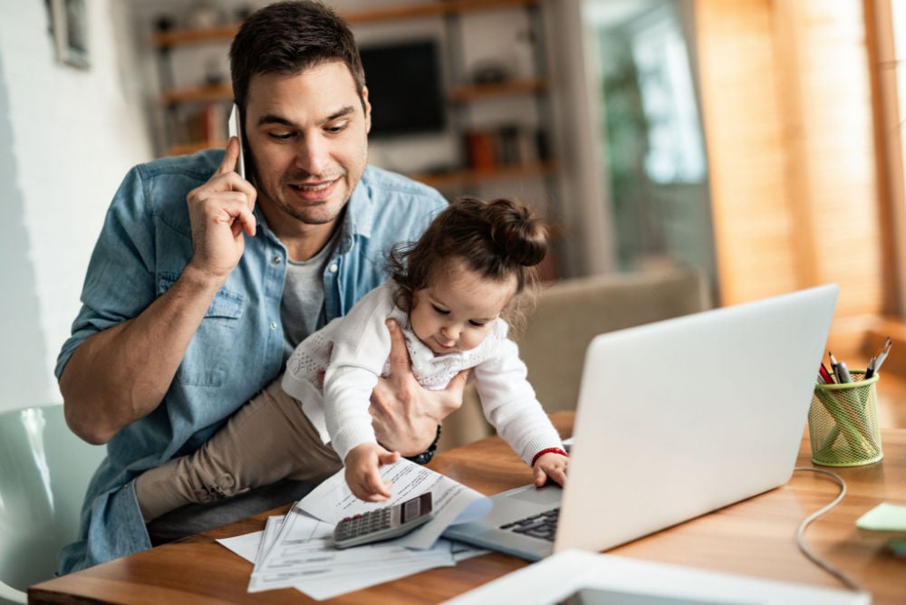 man working from home on a laptop while carrying his toddler daughter