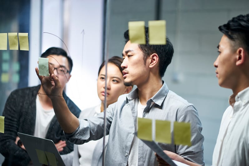 Three male and one female employee(s) in a strategy meeting. They are focusing on a glass screen with sticky notes pasted on it.