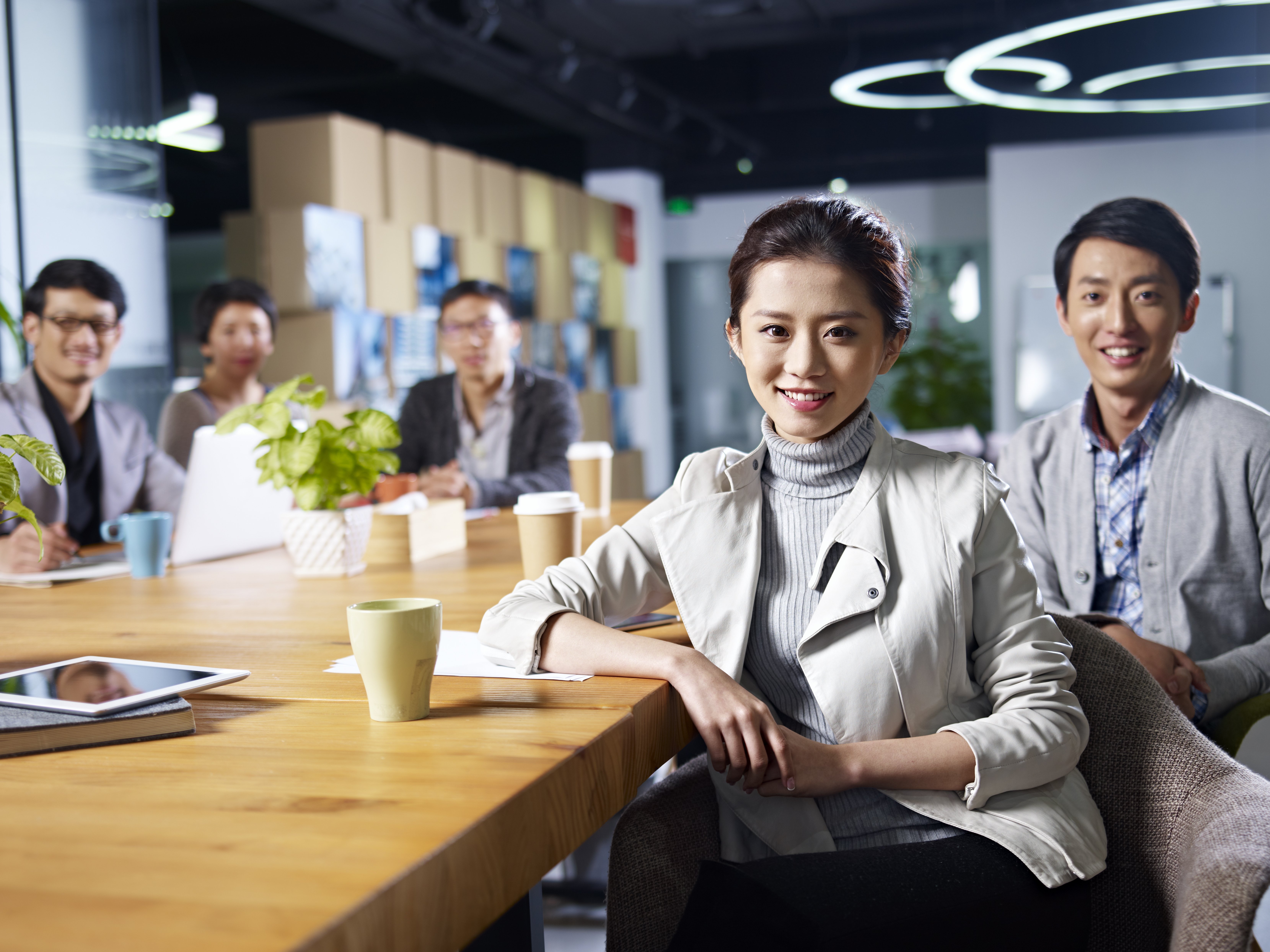 Woman seated at a long table in an office pantryand facing the camera and smiling. Behind her are four men also facing the camera and smilling. There are takeaway cups on the table and a potted plant.