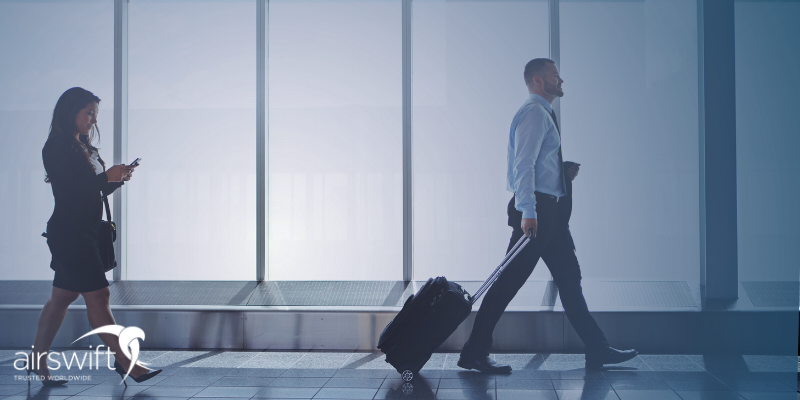 A smartly dressed man and woman walk through an airport. The man carries a suitcase, whereas the woman is texting on her phone