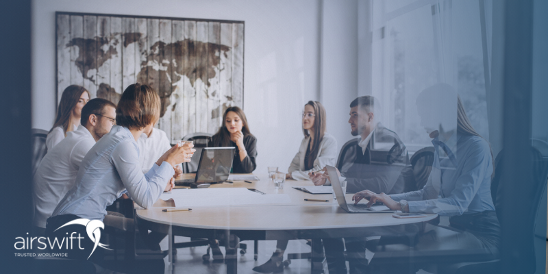a group of professionals sit around a desk in a glass office, having a meeting around a laptop