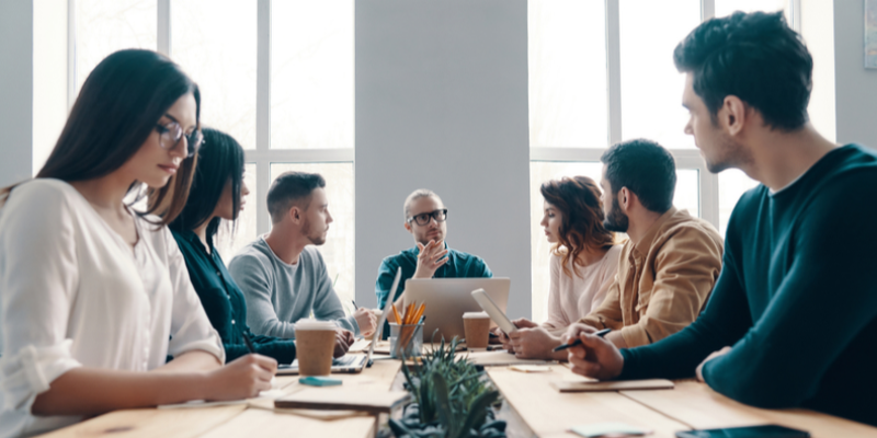 seven people sitting at a long wooden table in a middle of a work discussion